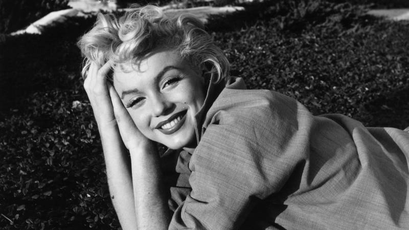 A black and white image of Marilyn Monroe, dressed casually in a loose shirt, reclining in the grass, smiling with tousled blonde hair.