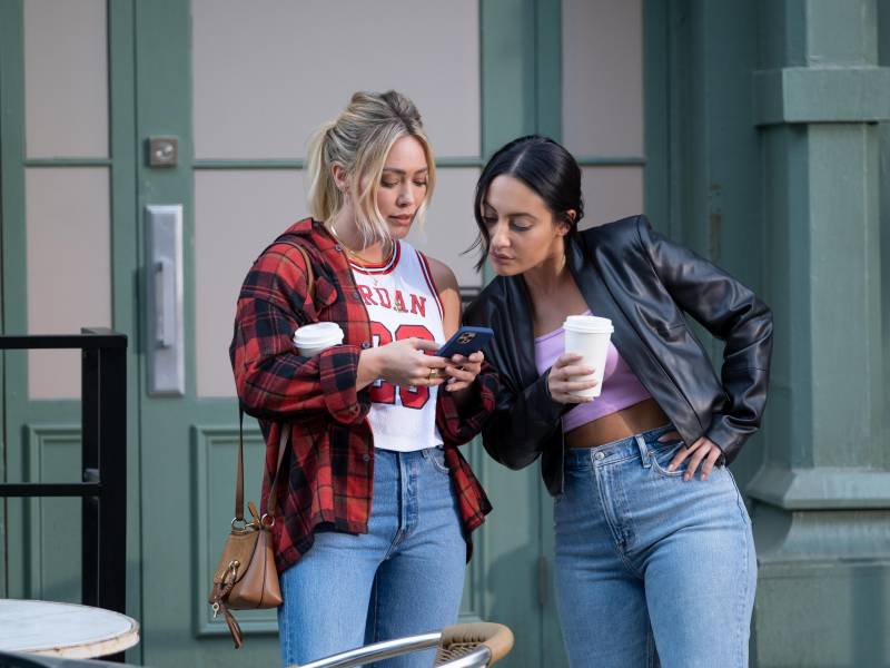 Two young women hold coffees and look at a phone together