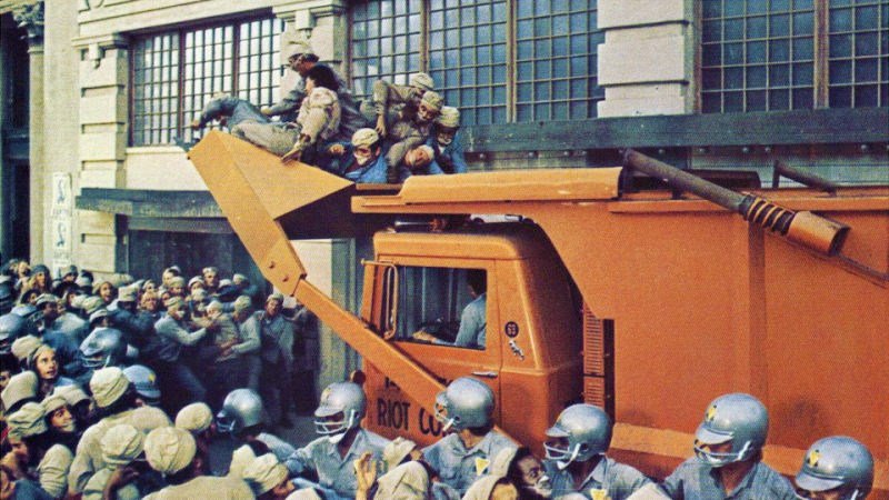An orange bulldozer scoops up a large group of people while crowds and police officers in helmets fight it out below.