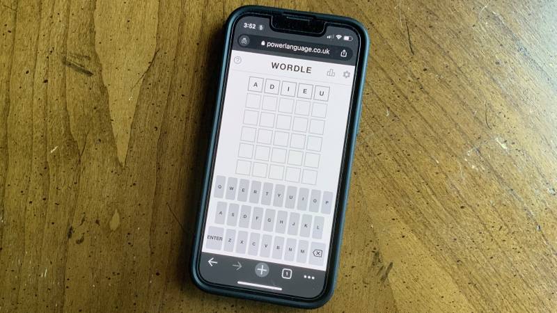 A cell phone lies on a wooden table. The screen shows a game of Wordle at play.