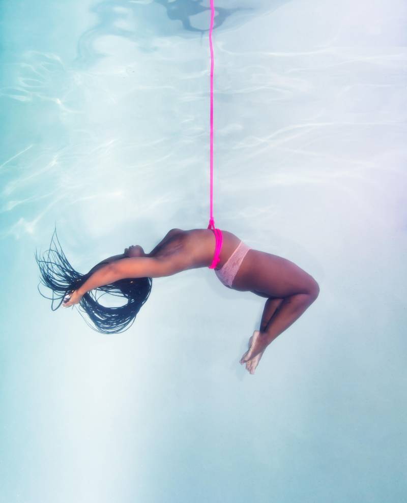A Black woman with long hair floats in the water, a bright pink rope wraps around her bare torso. The rope rises straight up towards the surface of the water.
