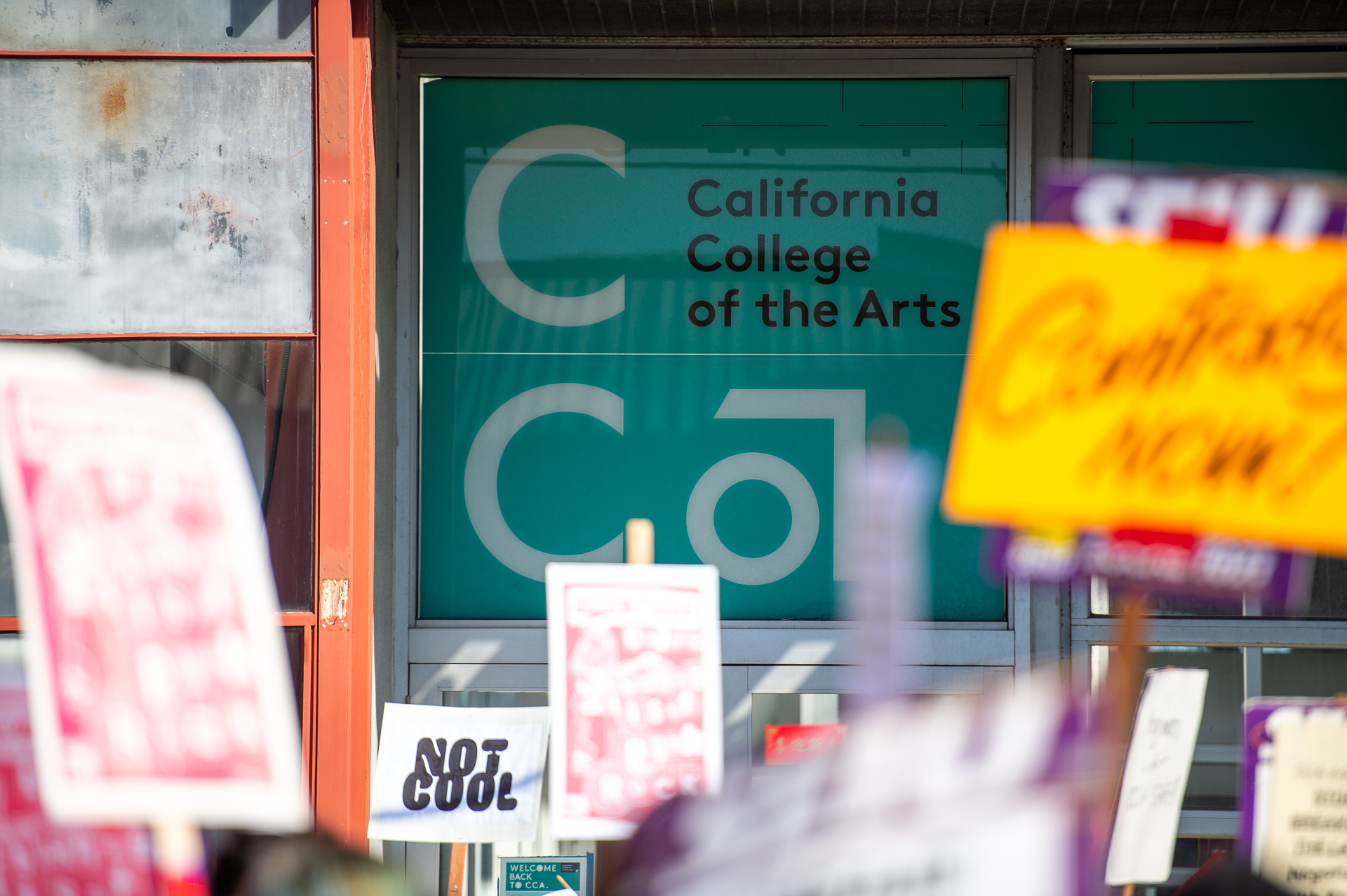 Picket signs read "not cool" and "contracts now" in front of a CCA logo