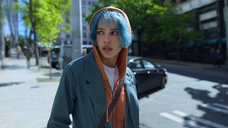 A worried looking woman with blue hair stands in the street, wearing an orange hoodie with the hood up underneath a grey suit jacket.