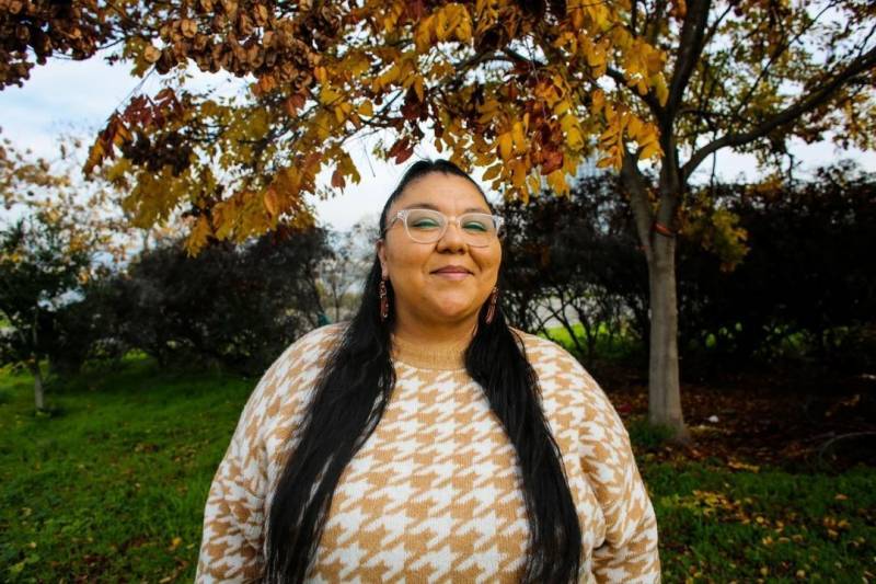 A portrait of Krea Gomez outside in front of fall leaves and green grass. She's smiling, wears clear rimmed glasses, tan houndstooth sweater and her long hair falls past her chest.