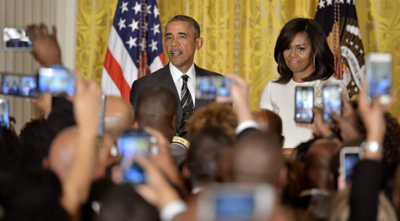 Barack and Michelle Obama stand on stage in front of a golden yellow curtain and American flag, while a packed audience holds up cell phones to take their photo.