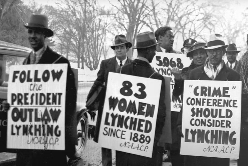 Black and white image of a gathering of protesters wearing sandwich boards with messages from the NAACP on them. The signs read: 'Follow the President. Outlaw Lynching'; '83 Women Lynched Since 1889'; 'Crime Conference Should Consider Lynching'.