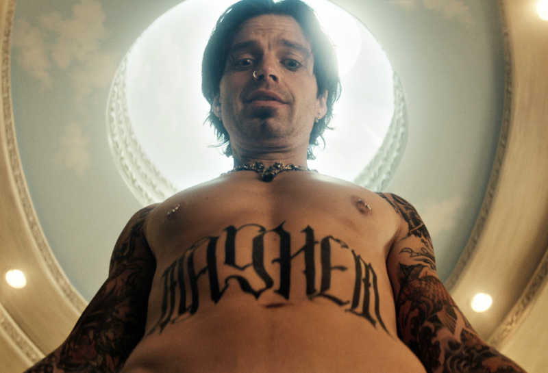 A naked man is seen from the waist up, as he stares down towards the camera. He has black hair, nipple piercings, heavily tattooed arms and the word 'MAYHEM' tattooed in block letters across his torso.