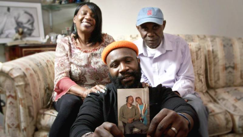 Karega, sitting on floor, holds a family photo to the camera, as his parents sit on a couch behind him. 