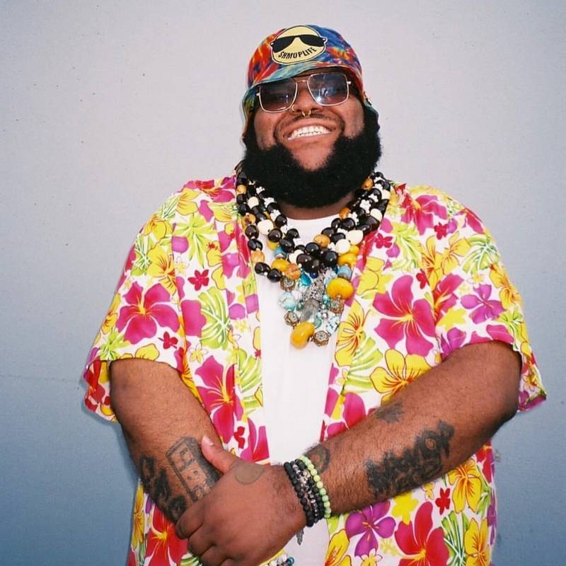 Oopz smiles at the camera, wearing a bright Hawiian shirt and a tye-die hat. He's got a chunky necklace and a few tattoos are visible on his forearms.