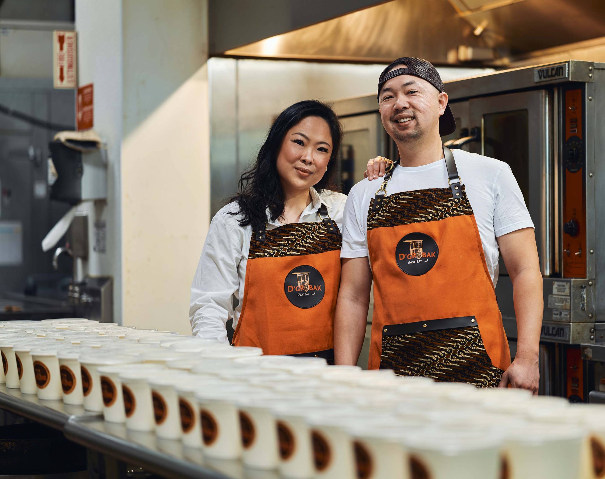 Two chefs in orange and brown aprons pose in their kitchen in front of many empty soup containers.