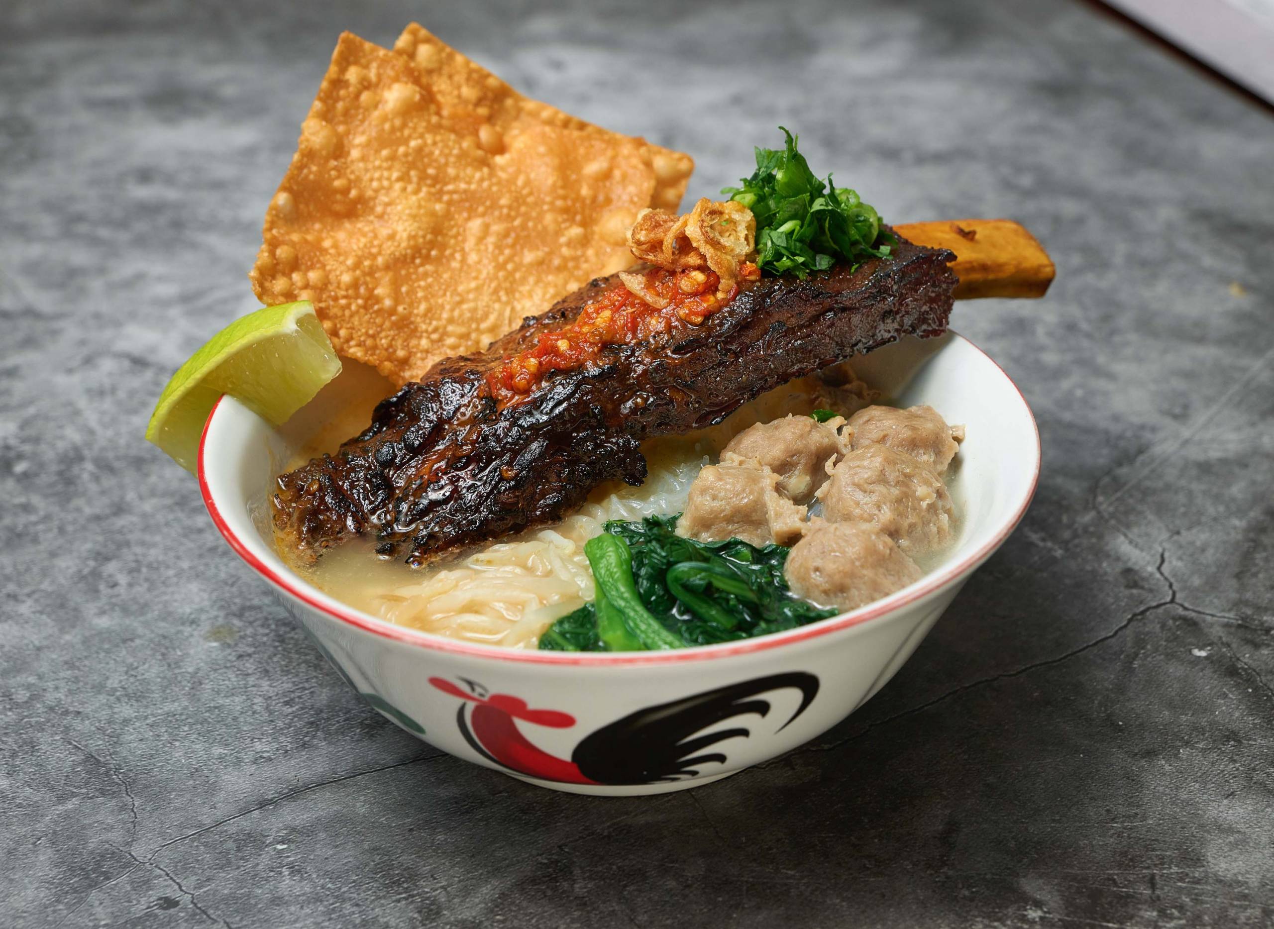 A bowl of bakso (Indonesian meatball noodle soup) topped with a whole beef short rib.
