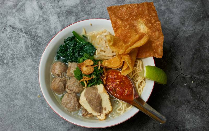 Overhead view of a bowl of noodle soup with meatballs and a spoonful of sambal.