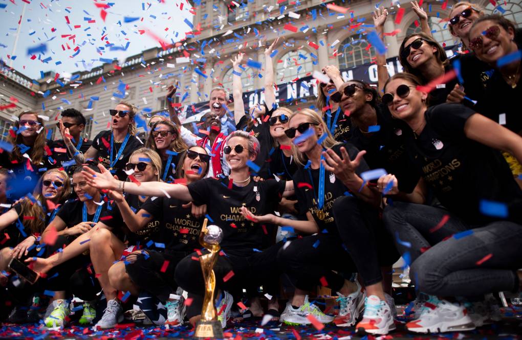 A group of smiling women, all wearing sunglasses, black outfits and matching white and orange sneakers, crouch together, smiling and waving their arms, as ticker tape falls all around them.