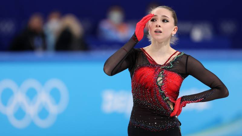 A figure skater, in black and red costume and red gloves pauses on the ice, one hand raised to her forehead.