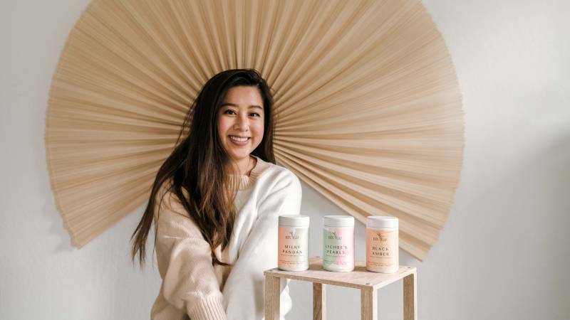 Kin Leaf Tea founder Violet Diana Nguyen poses with three canisters of her tea blends, in front of an ornamental fan.