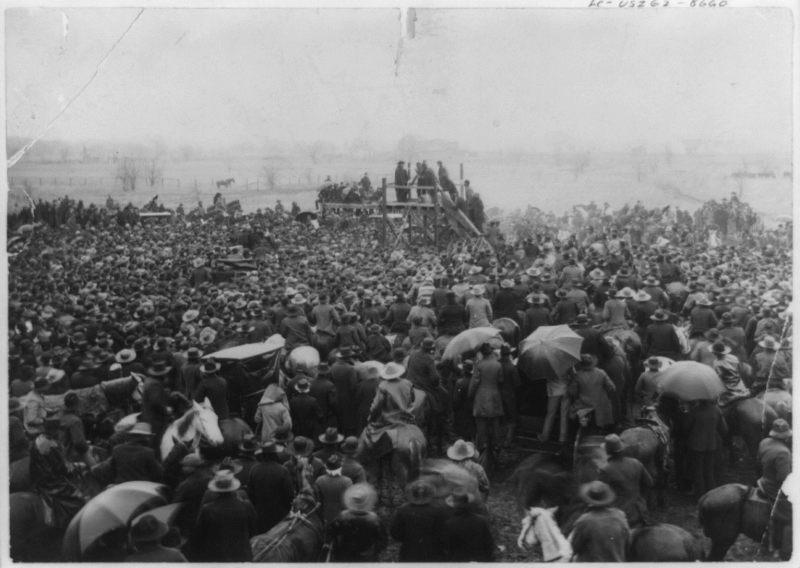 Hundreds of people gather in a field, some holding umbrellas, some sitting on horses. In the far distance, a ramshackle wooden platform with a few men standing on top.