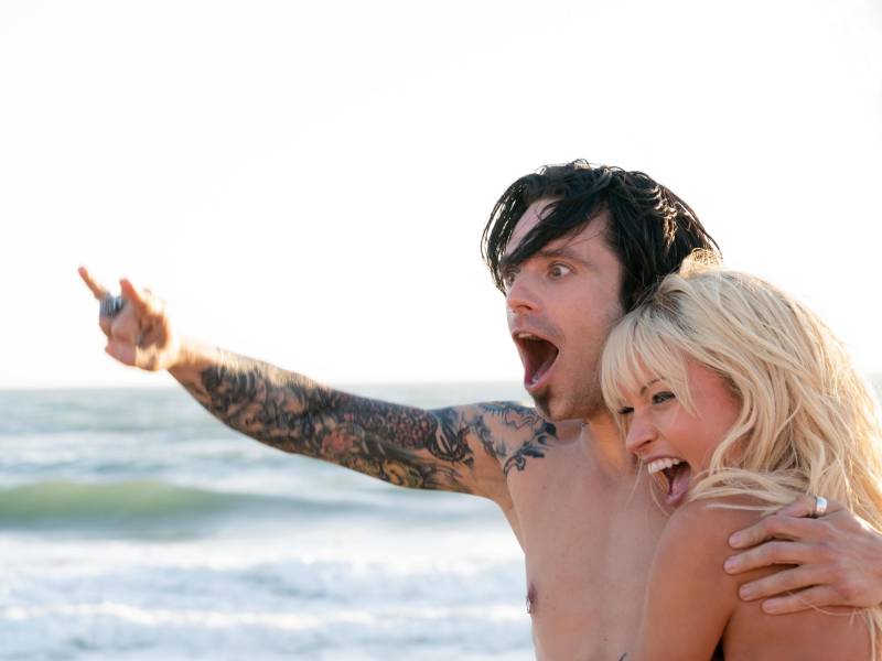 A tattooed man with one arm raised and another around a beautiful blonde woman stand before the ocean, celebrating.
