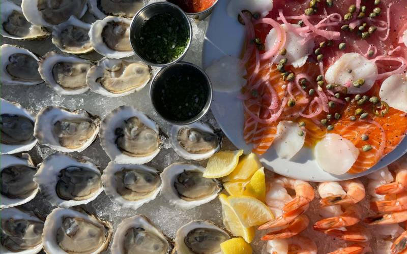 Overhead view of a seafood spread: raw oysters on the half shell over ice, shrimp cocktail, and fish crudo.