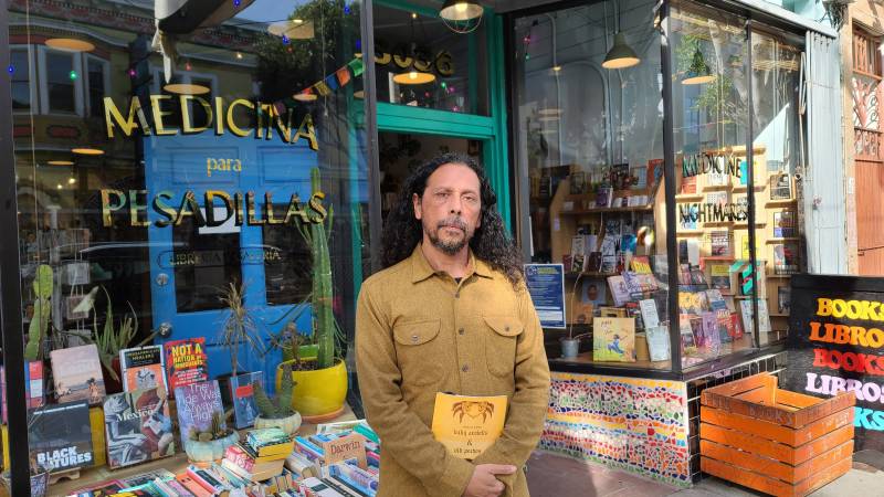 Josiah Luis Alderete stands in front of the windows of his bookstore, he wears a mustard shirt and holds his book of poems at his waist.