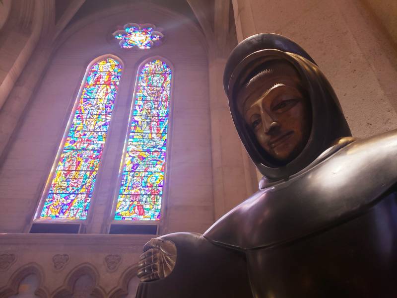 A bronze statue of a serene-looking man in monk-like robes, arms outstretched, stands before enormous, long, thin stained glass windows.