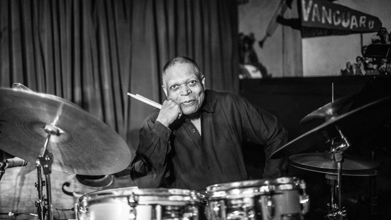 An African-American man sits at a drum kit in a jazz nightclub in a black and white photo.