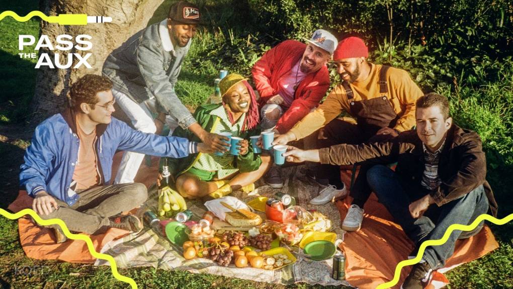 A band sits around a brightly colored picnic spread and clinks their mugs for a toast.