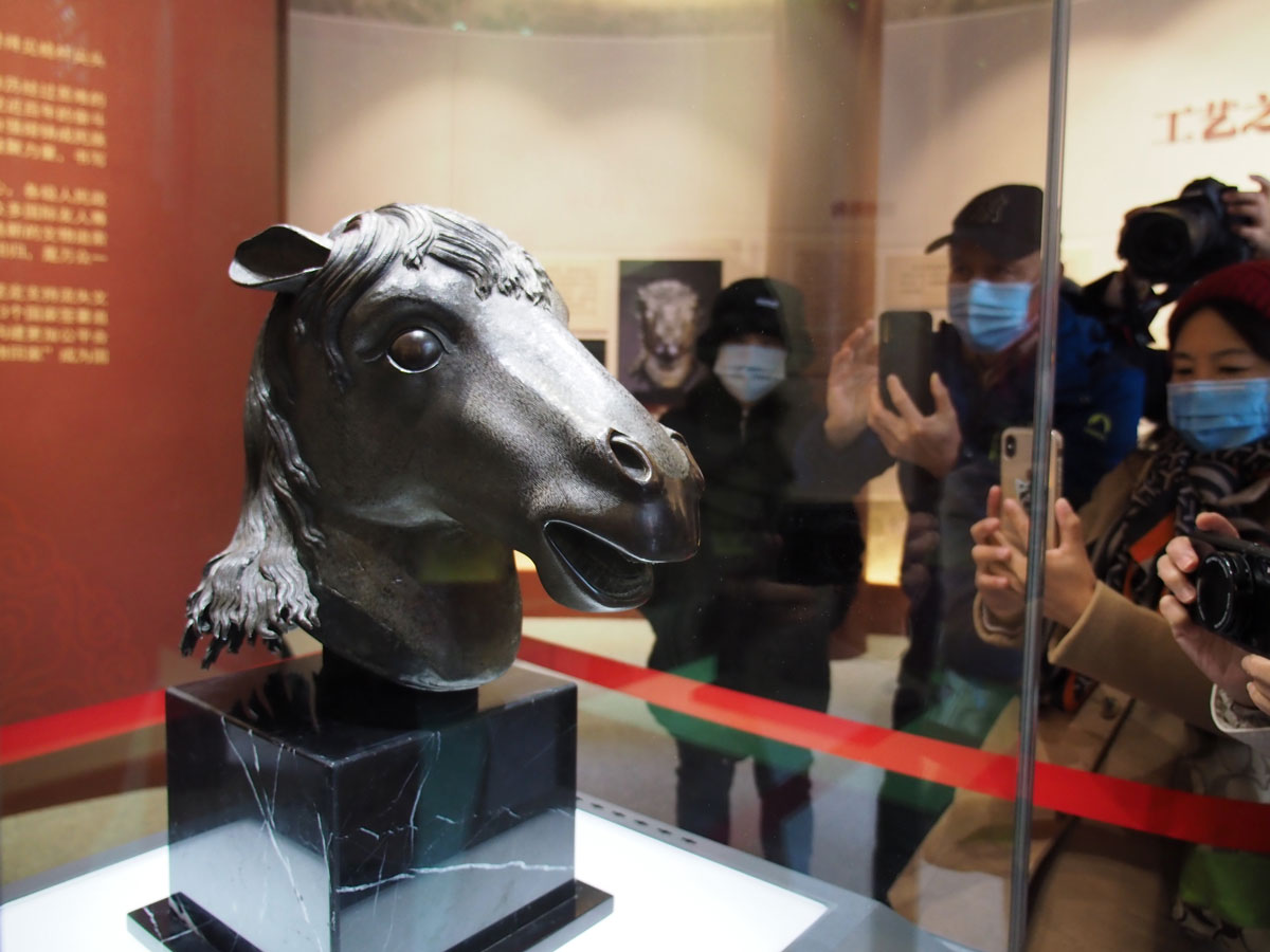 Horse head sculpture behind glass with people photographing it