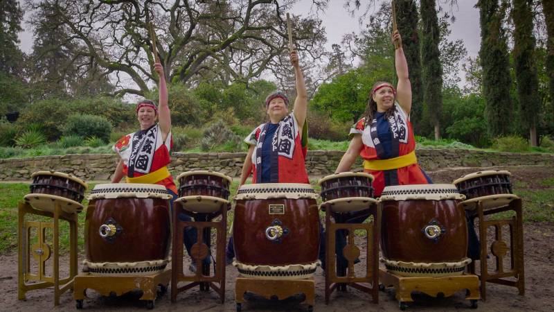 Three female taiko drummers raise their right hands while holding a drumstick.