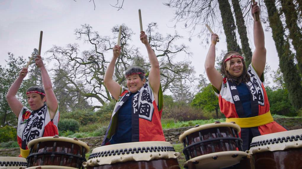 Three women wearing traditional taiko player attire raise their arms to srike big taiko drums with wooden sticks.