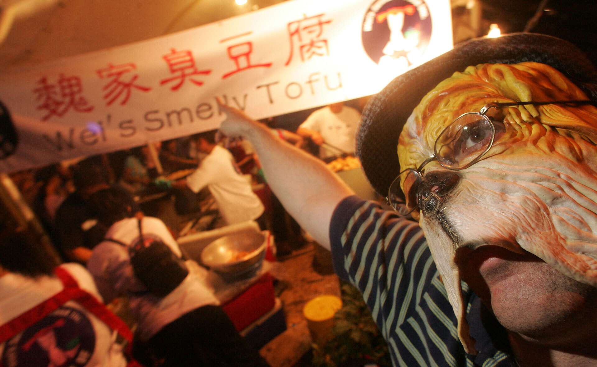 A man wearing a dog mask and glasses points to a street stall sign that reads, "Wei's Smelly Tofu."