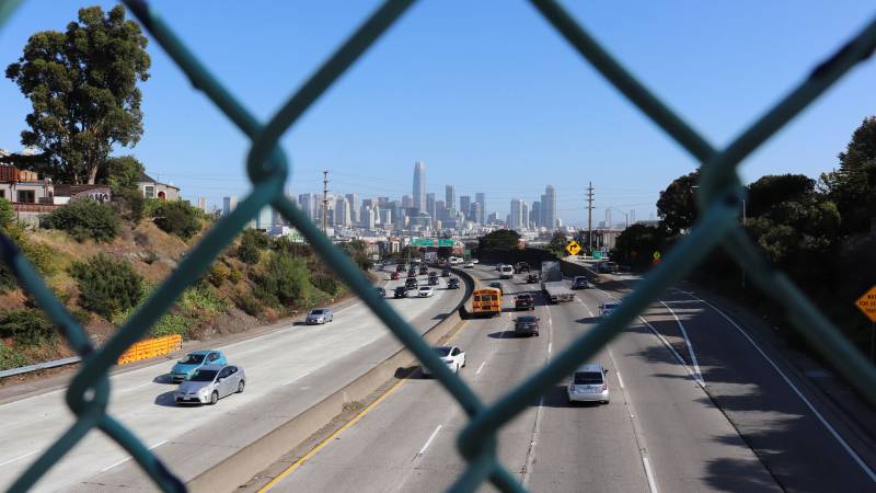 View of city on sunny day through chain-link fence over highway