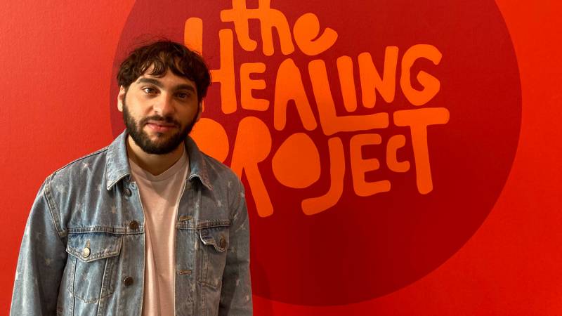 Man with dark hair and beard in denim jacket and white t-shirt standing in front of red wall art that says "The Healing Project" in a circle