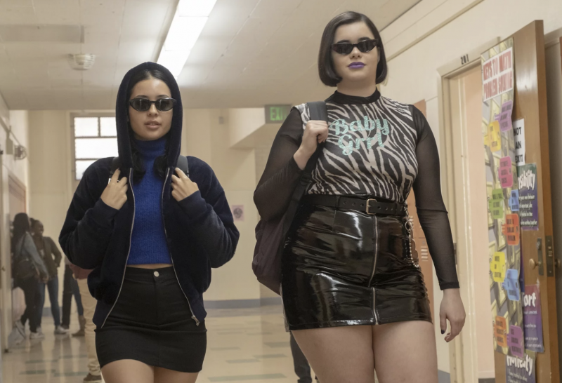 Two girls wearing very short black skirts, edgy tops and sunglasses walk confidently through a school hallway.