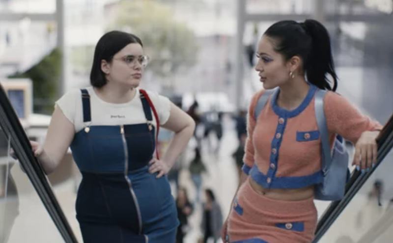 A girl wearing a skin-tight denim pinafore dress talks to a girl wearing a soft and skimpy pink and blue take on the classic Chanel suit.
