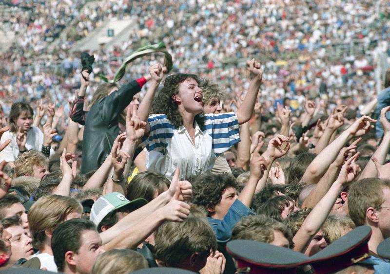 An enthusiastic crowd sings along at a stadium concert, including one young woman on someone's shoulders, arms raised in joy.