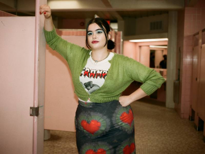 A young woman with black hair and bold green eyeshadow leans on a pink bathroom stall. She's wearing a green sweater with a black and white Cramps T-shirt underneath, paired with a skin tight black, red and grey skirt.
