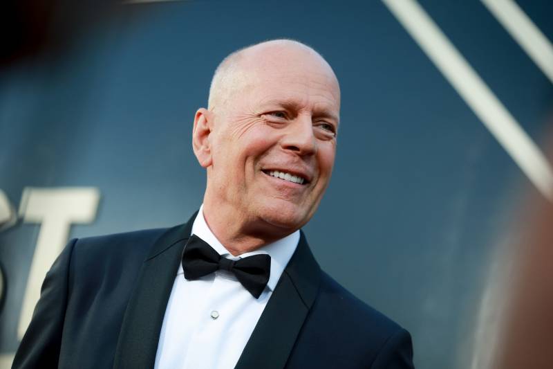 Bruce Willis, smiling broadly, wears a tuxedo at a red carpet event.