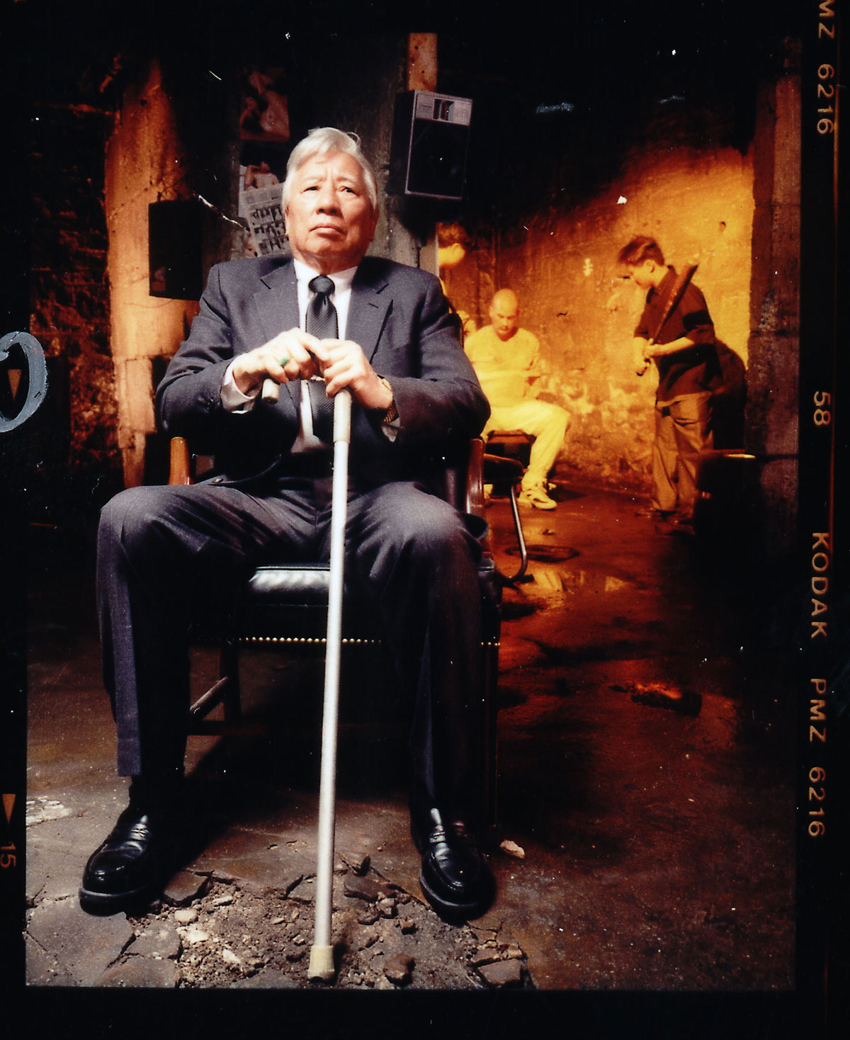 An older man in a suit sits with a cane in his hands, in the background the man is holding a bat in front of the man in the chair