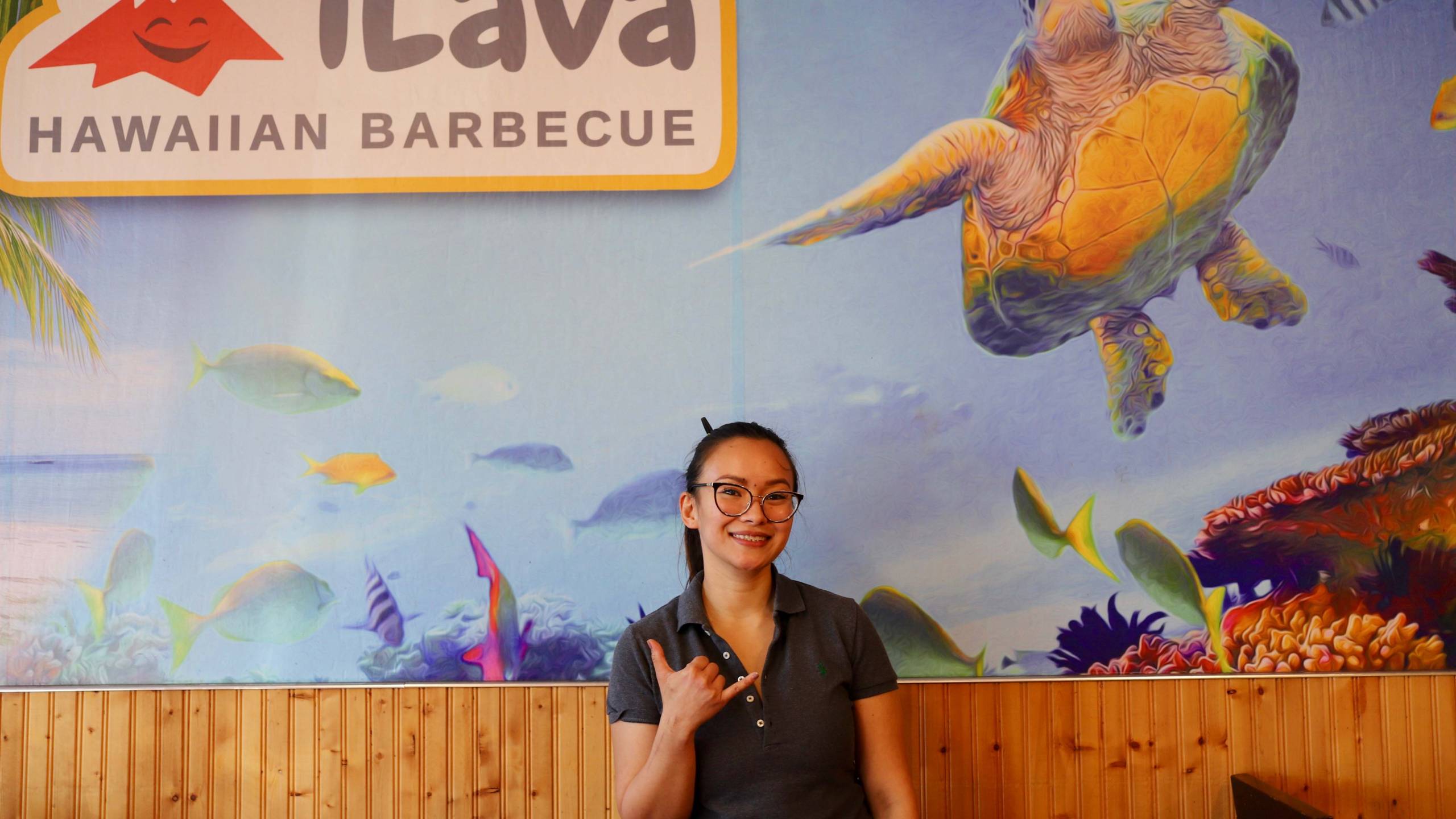iLava Hawaiian Barbecue owner Nicolle Jacinto makes a "hang loose" hand gesture in front of an ocean mural inside her East Oakland restaurant.