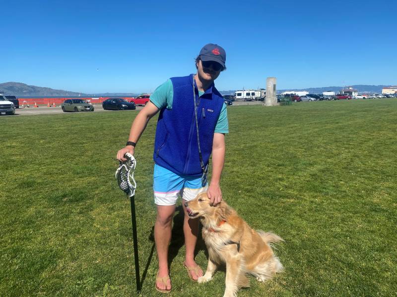 A guy in a blue vest, shorts, hat, playing on the Marina green with his dog
