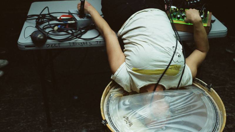 Performer lays with head down in a bass drum, hands reaching back to audio controls