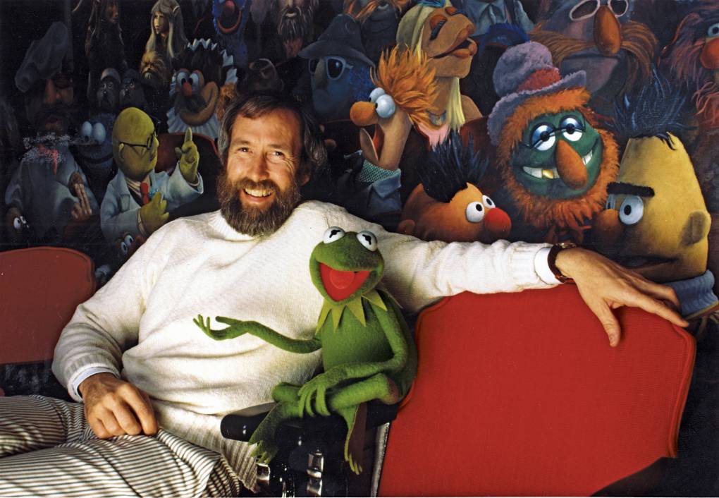 A white man with a beard sits with a green frog puppet, Kermit the Frog, in front of a colorful mural.