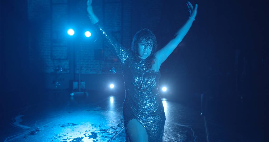A woman with curly hair in a sparkly dress dances in blue light