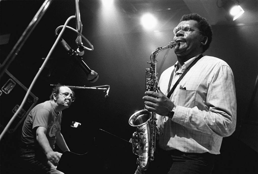 A middle-aged Black man plays the alto saxophone on a dimly lit stage