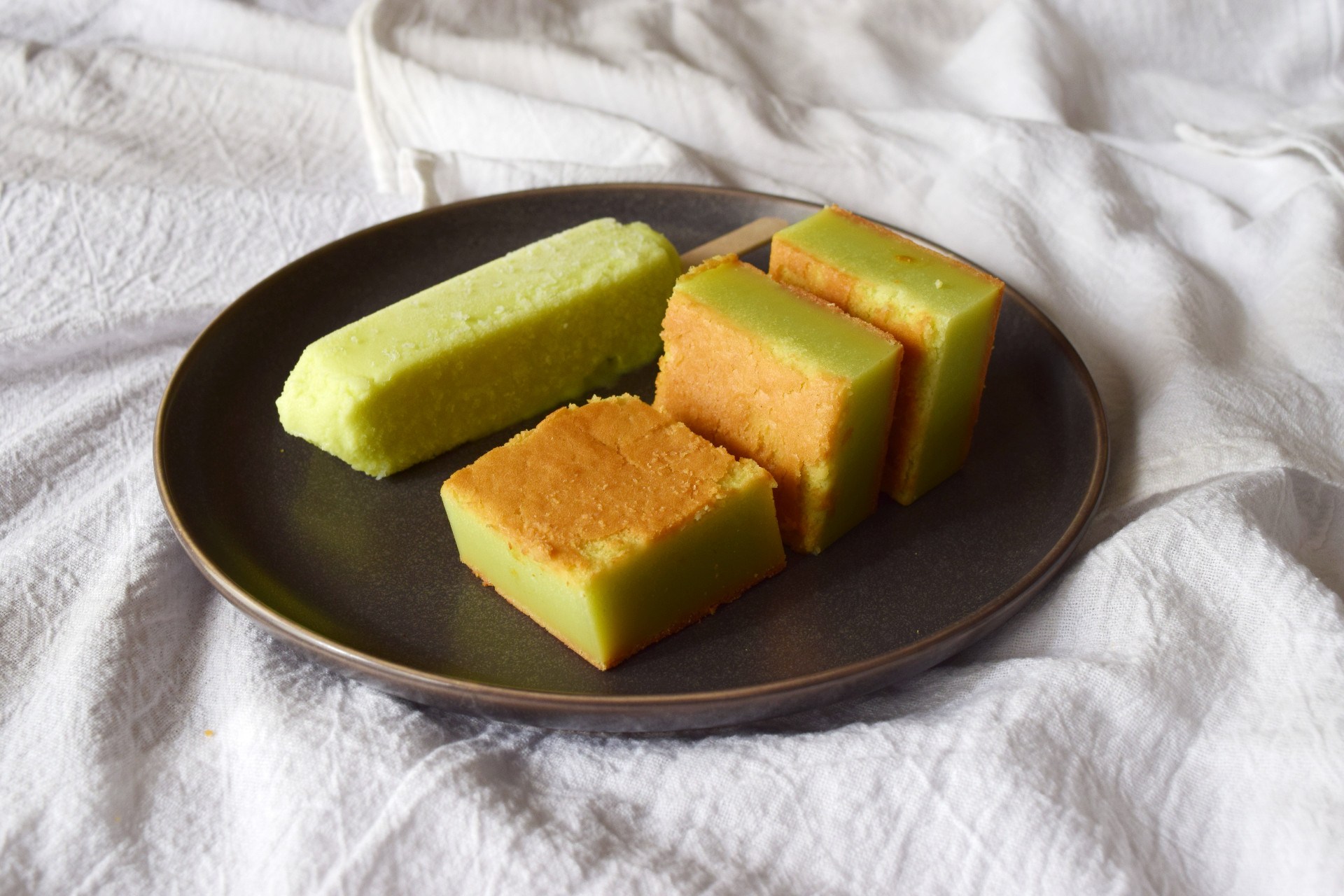 Three squares of melon-flavored mochi on a plate, next to a melon popsicle with the same pale green color.