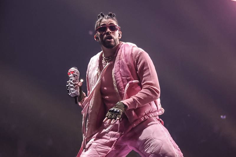 A man wearing head to toe pale pink athletic attire, biker gloves and red wrap-around sunglasses strikes a pose, mid-performance, microphone in hand.