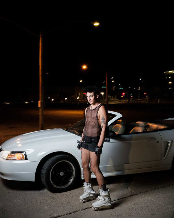 Andrea Abi-Karam is wearing a pair of white roller skates, some shorts and a black fishnet shirt while posing in front of a drop-top white Mustang