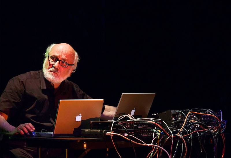 An elderly man at a string of laptops on stage.