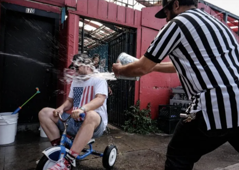 A referee in a black and white shirt splashes an adult competitor in a tricycle race with a bucket of water.