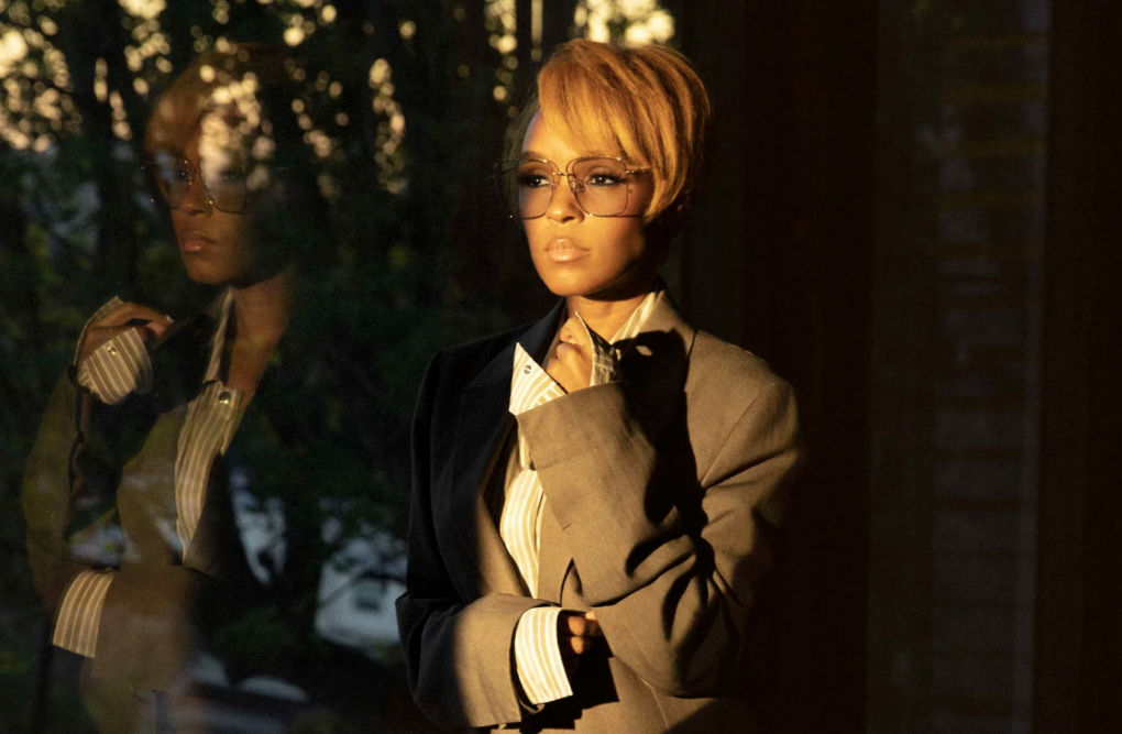 Janelle Monáe stands next to a window, her figure reflected in the glass, wearing a oversized man's suit and transparent sunglasses.